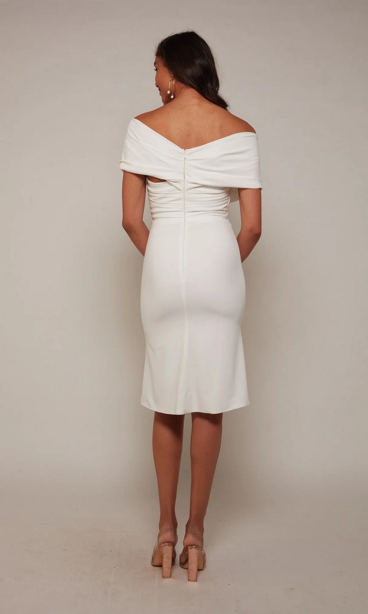 A woman stands facing away, wearing the Alyce 70023 Knee Length White Party Dress with nude heels against a plain, light background.
